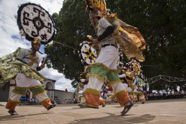 Teotiles dancing. In Teotitlan del Valle, the dance also highlights another contradiction. Fifty years ago, the town was very poor and much of the traditional weaving craft that had created part of its historical identity was no longer practiced. That poverty, reinforced by economic reforms and trade agreements that undermined Oaxaca's agricultural economy, forced many of the town's residents to leave. They became migrants, first within Mexico and then across the border into the United States. As remittances began arriving to support the families left behind, expatriates also provided money to buy materials for weaving. With the influx of tourists anxious to buy rugs in traditional Zapotec designs, weaving workshops were reestablished. Migrants saved money to buy the materials for the elaborate clothing and headdresses needed for the Dance of the Feather. Some returned home to fulfill the three-year commitment required of those wanting to perform the dance.  Teotitlan has a complicated relationship with migration. The remittances helped to revitalize the town. The workshops now weave not just for tourists, but also for museums in the U.S. But how easy is it to keep a culture in an economy that still depends heavily on the willingness of people to leave home to seek work elsewhere? “This dance is also a strategy for defense against what they felt were negative influences of the modern world, against the consequences of migration, against the loss of moral values and customs,” Hernandez emphasizes.