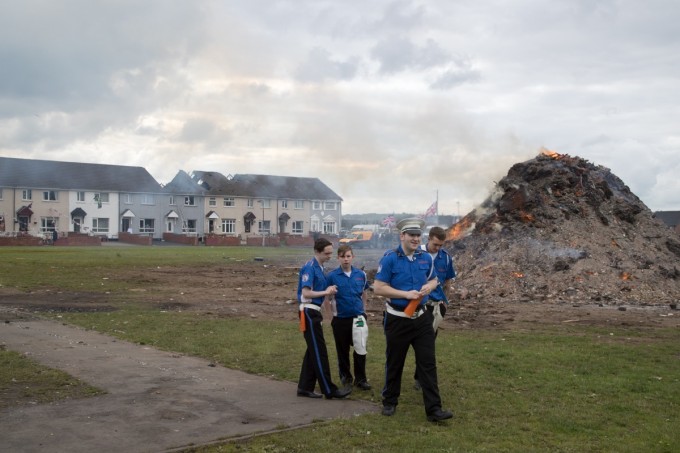 Young band members visit the smoldering remains of the Shankill bonfire before parading through the city. The damaged row houses are in the background.