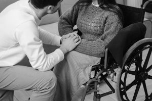 A black and white image of a person in a wheelchair holding hands with another person sitting down in front of them.
