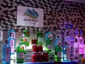 A backlit shelf of liquor bottles, in front of the tiled backsplash of Club Q's bar, features a political flyer for Jared Polis, who would become the first openly gay governor in the United States.